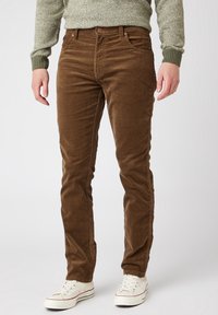 Man som bär bruna kordbyxor, grönstickad tröja och vita tygskor står mot en enkel bakgrund.