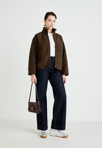 Woman wearing a brown fleece jacket, white shirt, dark wide-leg jeans, silver sneakers, holding a brown handbag against a plain white background.