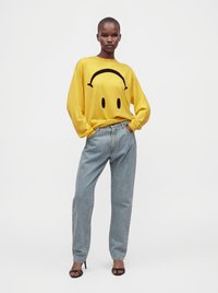 Femme portant un sweat-shirt jaune avec un visage triste noir, un jean bleu clair et des sandales à talons noirs, debout les mains dans les poches.