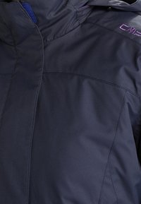 Veste imperméable bleu marine avec fermeture éclair frontale, capuche ajustable et détail de logo violet. Présente une texture lisse et mate ainsi qu'un design épuré.
