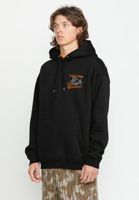 Czarna bluza z kapturem i kieszenią kangurzą, z sznurkiem do regulacji kaptura oraz pomarańczowym haftowanym logo. Teksturowana tkanina, luźny krój. Połączona z spodniami w kamuflaż.