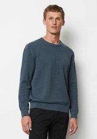 Blauer, texturierter Pullover mit rundem Halsausschnitt sowie gerippten Bündchen und Saum, mit einem kleinen Logo auf der Brust. Getragen mit dunklen Hosen.
