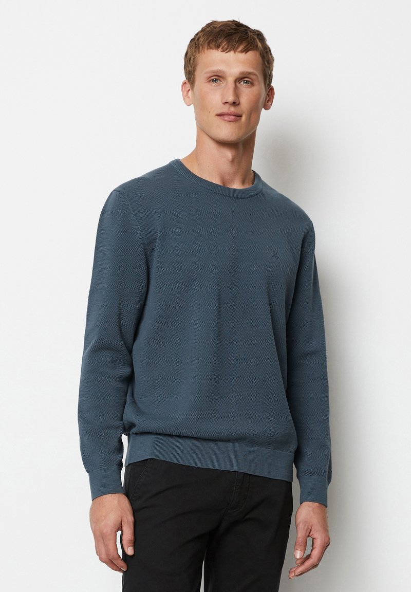 Blauer, texturierter Pullover mit rundem Halsausschnitt sowie gerippten Bündchen und Saum, mit einem kleinen Logo auf der Brust. Getragen mit dunklen Hosen.