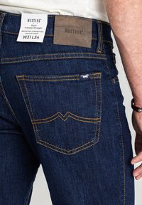 Ciemnoniebieskie jeansy z denimu o prostym kroju nogawki, z żółtym przeszyciem i brązową skórzaną naszywką marki na tylnej części pasa.