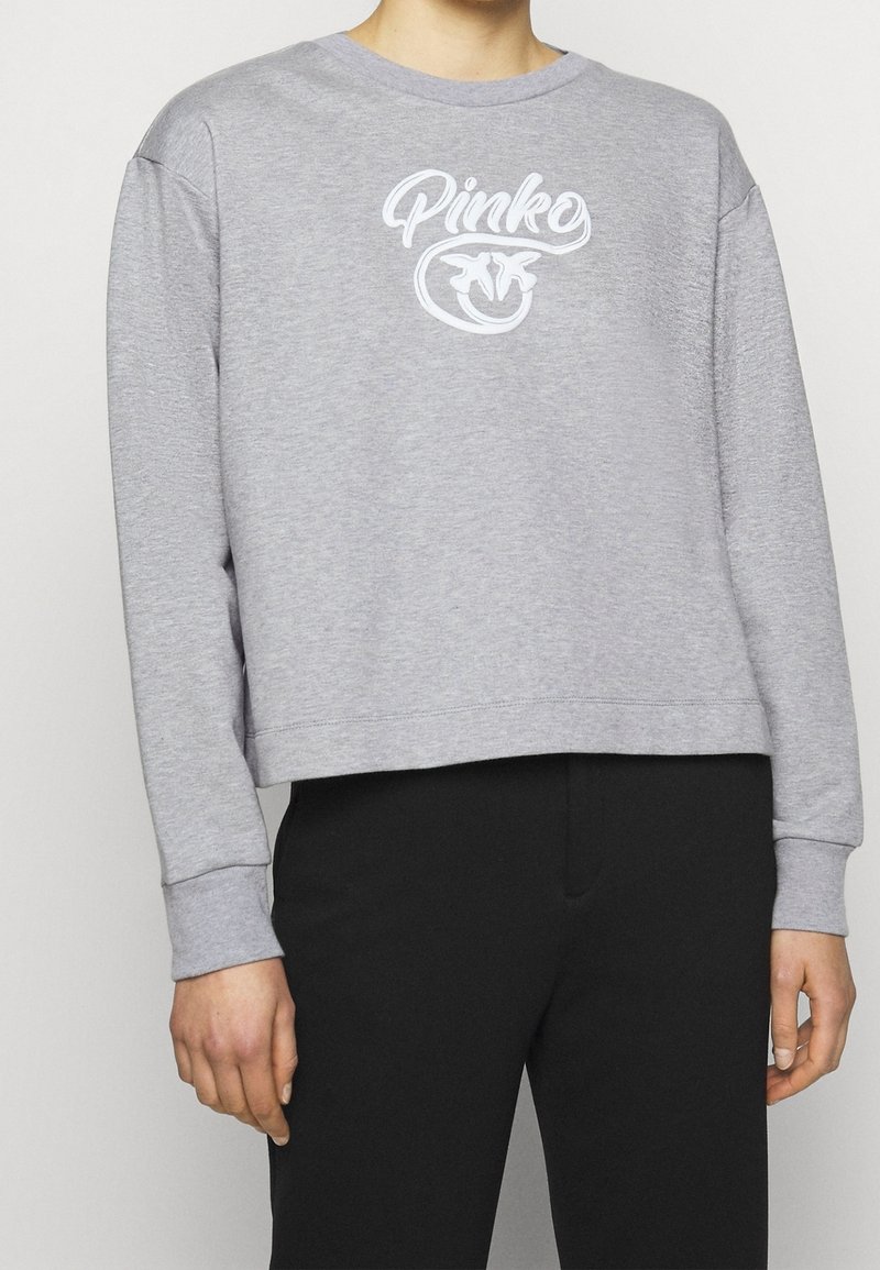 Personne portant un sweat-shirt gris clair avec un texte et un logo blancs "Pinko" sur le devant, associé à un pantalon noir, visible du cou aux hanches.