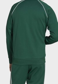 Grön sportjacka i ett slätt tyg med vita kantlinjer längs axlarna. Har en hög krage och ribbad mudd, i kombination med matchande byxor.