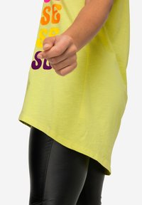 La maglietta gialla oversize presenta un testo colorato in un font ondulato. La parte superiore ha un orlo arrotondato, abbinata a leggings neri lucidi.