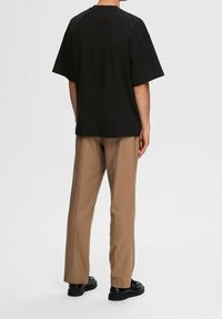 Persona con una camiseta negra holgada, pantalones rectos beige y zapatos negros, de pie contra un fondo claro liso, mirando hacia atrás.