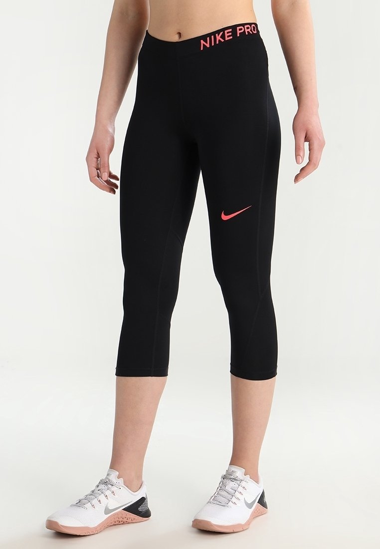 Czarne getry Nike Pro do połowy łydki z różowym logo na pasku i przodzie. Gładki, elastyczny materiał, dopasowany krój. Noszone z białymi i szarymi butami sportowymi.