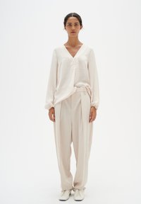 Helles beige, strukturiertes Blusenshirt mit V-Ausschnitt; kombiniert mit hochtaillierten, locker sitzenden Hosen in passender Farbe. Weiße Schnürschuhe runden den Look ab.