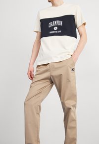 Beige och marinblå färgblockerad t-shirt med tryckt logotyp "Champion" och "Rochester 1919", parat med ljusbeige raka byxor.