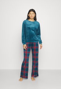 Haut en velours teal à manches longues associé à un pantalon évasé à carreaux dans des tons de vert, rouge et bleu. Texture douce et coupe décontractée.