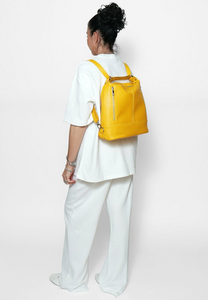 Geel leren rugzak met dubbele schouderbanden, twee ritsvakjes aan de voorkant en een gestructureerde vorm, gedragen tegen een simpele achtergrond.