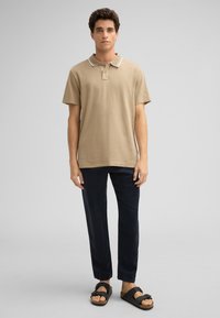 Beige Poloshirt mit weißem Kragen, kombiniert mit dunkelblauen Hosen und schwarzen Sandalen. Der Stoff wirkt glatt; lässiger Schnitt.