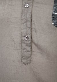 Tessuto grigio chiaro con una texture morbida, caratterizzato da una patta con bottoni e una tasca decorata con paillettes sul lato sinistro.