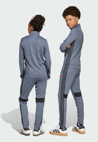 Grauer Sportanzug mit schwarzen Streifen an den Seiten; beinhaltet einen Reißverschlusskragen und schwarze Akzente an der Hose. Die Schuhe haben ein weiß-schwarzes Design.