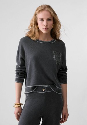 Donna con lunghi capelli biondi indossa un maglione e pantaloni coordinati grigio scuro con dettagli cuciti bianchi, orecchini a cerchio dorati e bracciali dorati spessi.