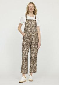 Salopettes en imprimé léopard dans un tissu marron et beige, avec des bretelles réglables, une poche avant et des jambes larges, assorties à un t-shirt blanc et des baskets.