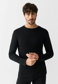 Black long-sleeve shirt made of ribbed fabric, featuring a round neck and fitted design. Model is holding hands in front.