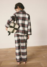 Barn i ternede pyjamas holder en indpakket gave bag ryggen og står barfodet på trægulv foran en neutral væg.
