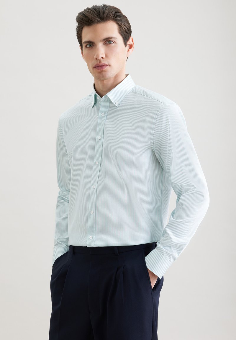 Jeune homme debout portant une chemise à rayures bleues claires à boutons et un pantalon foncé, sur un fond uni clair.