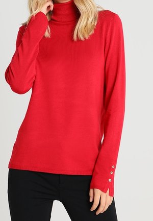 Femme portant un pull à col roulé rouge à manches longues avec des détails de boutons sur le poignet et un pantalon noir.