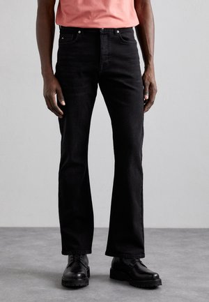 Mand iført sorte jeans med lige ben, sorte snørestøvler og en lakserød t-shirt mod en ensfarvet grå baggrund.