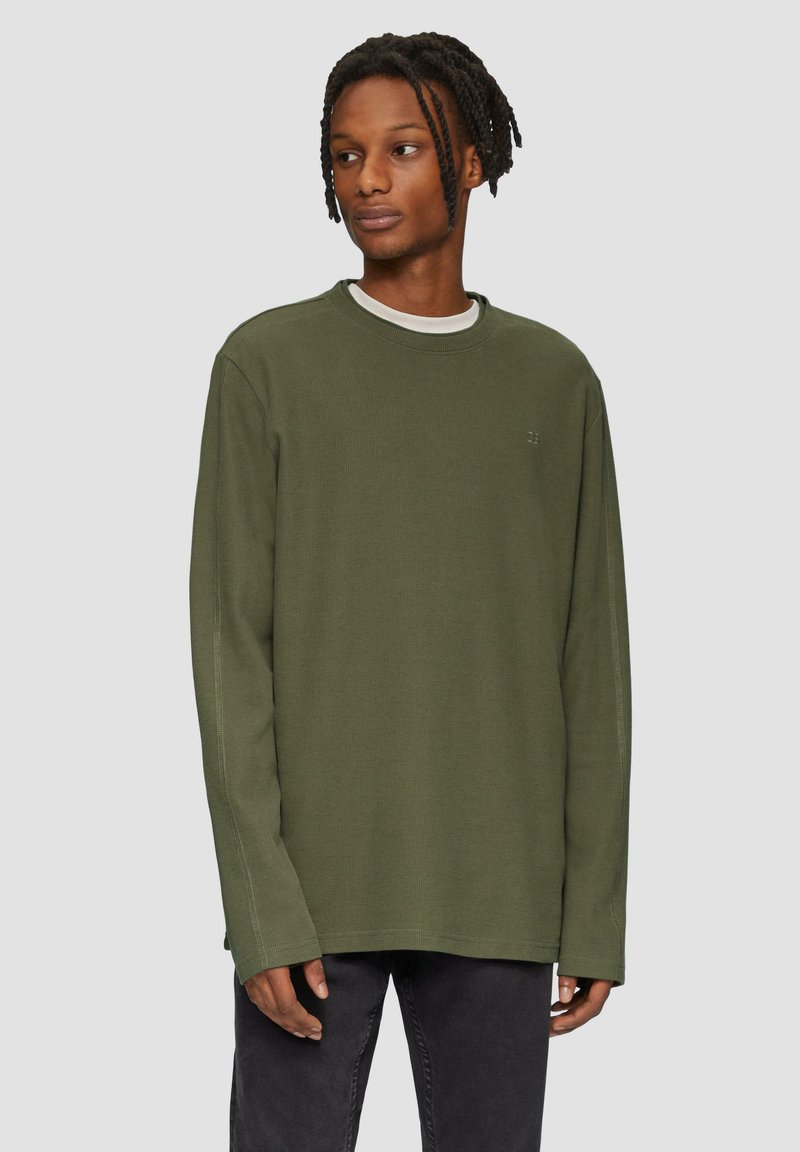 Langärmliges, olivgrünes, geripptes Shirt mit Rundhalsausschnitt und gesticktem Logo auf der Brust, kombiniert mit dunklen Hosen. Weiche Textur.