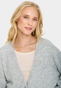 Heled hall kardigan, millel on V-kaeluse disain ja pehme tekstuur, varustatud nööbistamisega, seljas kreemikat värvi pluusi peal.