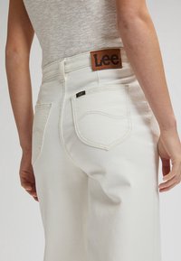 Personne portant un jean blanc taille haute avec un patch logo Lee et des poches arrière cousues, associé à un top beige sans manches.