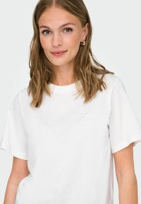 Weißes Baumwoll-T-Shirt mit Rundhalsausschnitt, kurzen Ärmeln und einer kleinen gelben gestickten Gänseblümchen auf der linken Brust. Glatte Textur.
