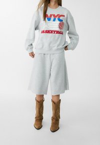 Harmaa huppari, jossa on "NYC BASKETBALL" -grafiikka, yhdistettynä sopiviin harmaisiin shortseihin ja ruskeisiin anteekka-suede cowboy-kengiin, joissa on terävä kärki.
