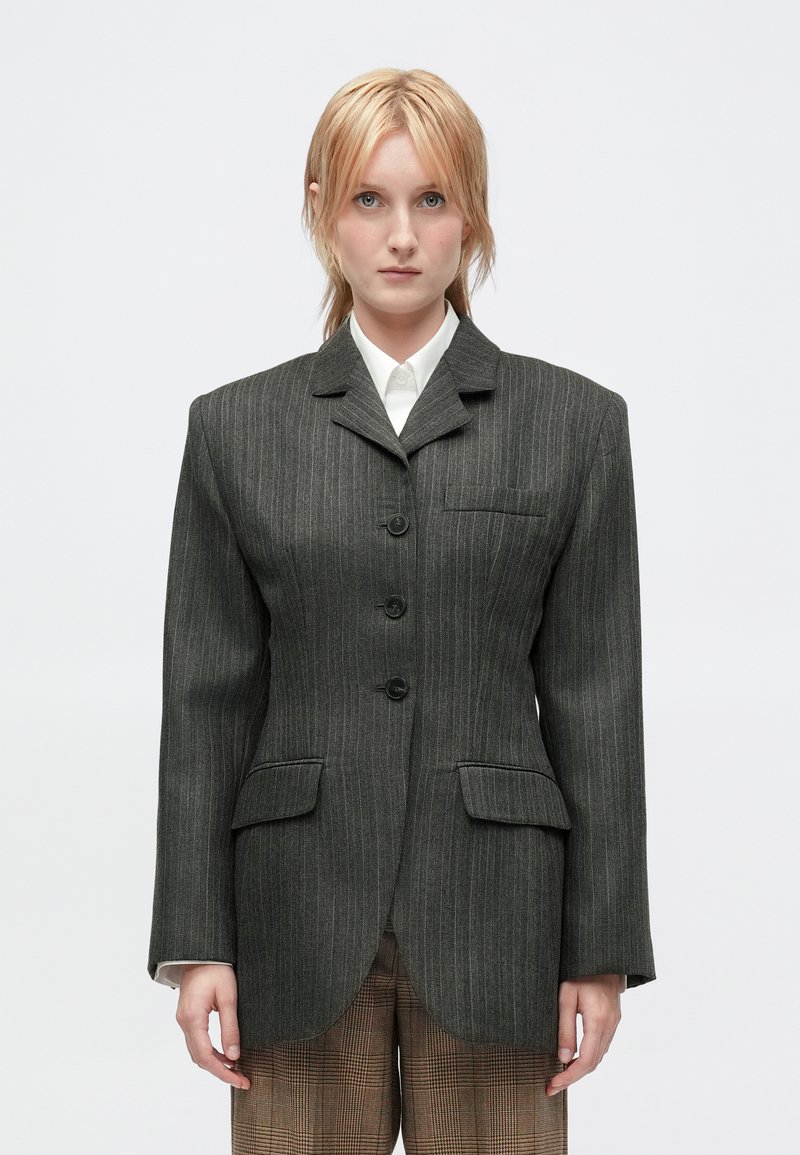 Een persoon met kort blond haar draagt een donkergrijze gestreepte blazer over een wit shirt en bruine geruite broek tegen een effen lichte achtergrond.