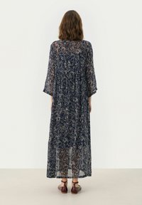 Rochie lungă, fluidă, de culoare bleumarin, cu un model floral, dintr-un material transparent, cu mâneci lungi până la cot și un design cu fuste în straturi. Purtați cu sandale cu barete maro.