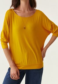 Lockere, leuchtend gelbe Bluse aus weichem Stoff mit weitem Ausschnitt und Dreiviertelärmeln, kombiniert mit dunkelblauen Jeans. Sichtbare Goldkette.
