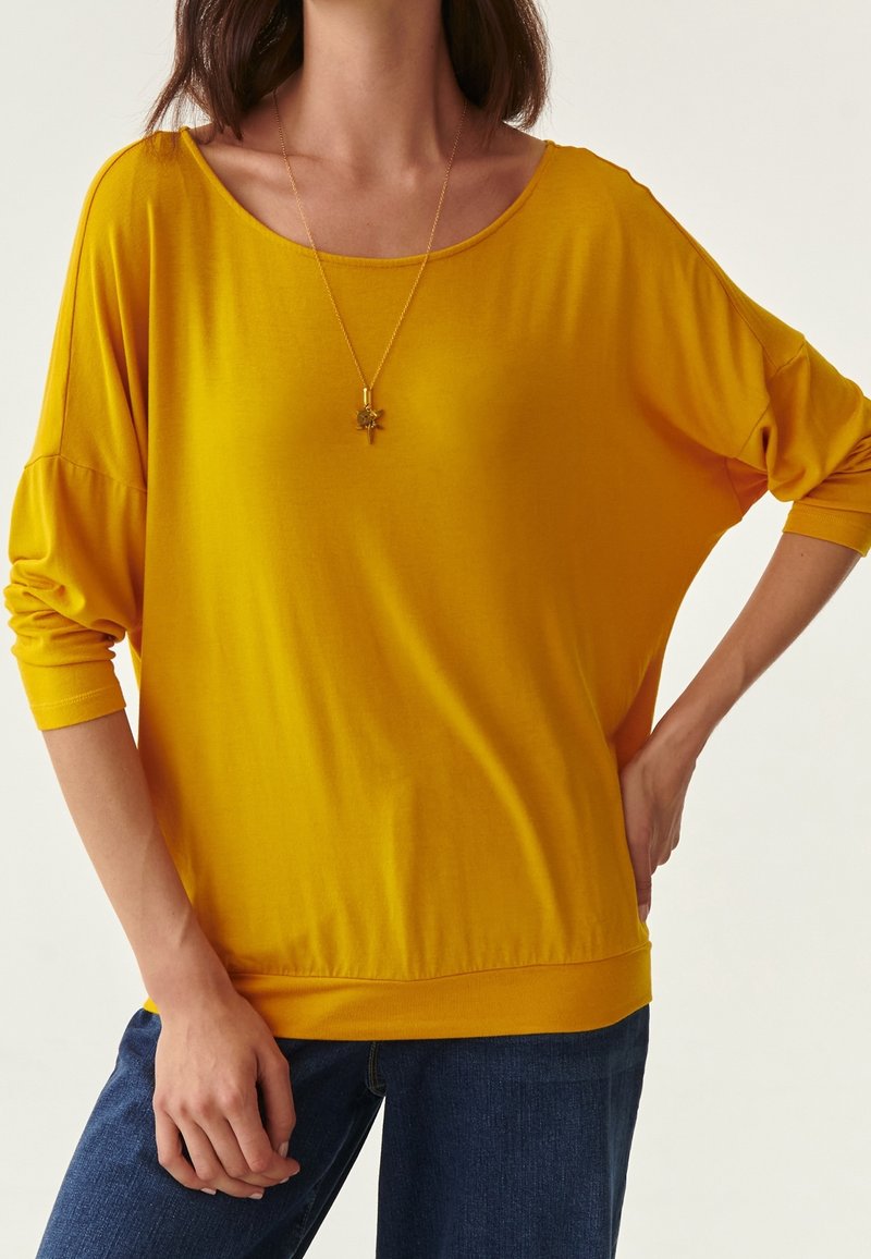 Lockere, leuchtend gelbe Bluse aus weichem Stoff mit weitem Ausschnitt und Dreiviertelärmeln, kombiniert mit dunkelblauen Jeans. Sichtbare Goldkette.