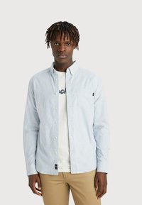 Lys blå og hvid stribet skjorte med knapper, lavet af bomuldsstof, lange ærmer, forlommer, båret over en hvid t-shirt, parret med beige bukser.