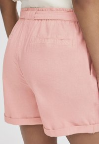Lys rosa shorts laget av et strukturert stoff, med en samlet midje, sidelommer og rullede mansjetter. To baklommer synlige.