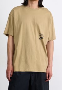 Camiseta de algodón beige con manga corta, cuello redondo y un logotipo impreso en negro y texto en el lado izquierdo del pecho. Textura suave.