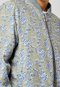 Blouson aviateur fleuri vert olive avec motifs floraux bleus et blancs. Présente un col côtelé, une fermeture éclair et un tissu texturé.