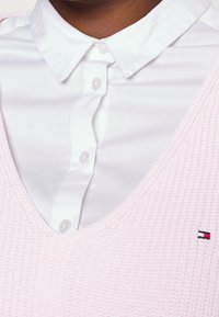Veste en tricot rose clair avec un col en V superposée à une chemise blanche à boutons, arborant un petit logo sur le côté gauche.