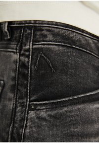 Lähikuva haalistuneista mustista farkuista, joissa näkyy ompeleita, niitti ja osa valkoisesta paidasta, joka on työnnetty vyötärön yläpuolelle.