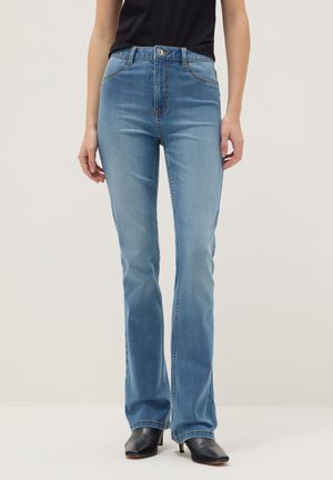 Femme portant un jean bootcut bleu clair avec poches avant et un haut noir, debout les mains détendues le long du corps.
