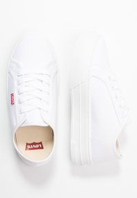 Baskets en toile blanche avec lacets plats, bout rond et semelle en caoutchouc surélevée. Présentent un petit logo Levi's rouge sur le côté.