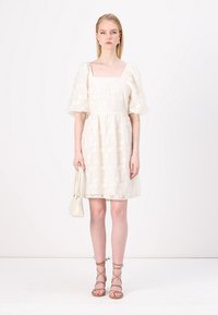Weißes kurzes Kleid mit Puffärmeln, strukturierter Stoff, quadratischer Ausschnitt, tailliert und mit weitem Rock; kombiniert mit nude-farbenen Sandalen mit Riemchen und einer kleinen weißen Tasche.