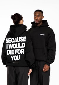 Sudaderas negras con corte oversize; el texto en blanco en la parte posterior dice "PORQUE MORIRÍA POR TI." Pequeño logo en el lado izquierdo del pecho. Textura de tejido suave.