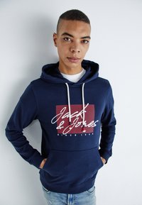 Felpa blu con cappuccio e cordini, con un design del logo bianco e rosso sul petto, polsini a coste e una tasca frontale.