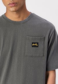 Grey T-shirt featuring a round neck, short sleeves, and a left chest pocket with a black label displaying "Stan Ray" in gold lettering.