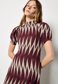 Robe tricotée avec un col haut, des manches bouffantes et un motif en zigzag dans des nuances de bordeaux et beige. Texture lisse avec une silhouette ajustée.