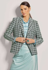 Grüner und weiß gemusterter Blazer mit scharfen Revers, kombiniert mit einer hellblauen Satinbluse und einem Rock. Auffällige Akzente setzen ein großer türkisfarbener Ring.